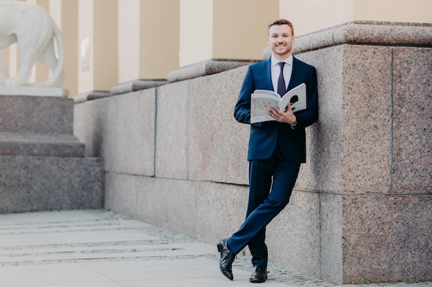 La foto de un exitoso jefe masculino se para con las piernas cruzadas sostiene una revista de negocios que lee la información necesaria se prepara para la presentación frente a sus colegas se encuentra cerca del edificio de oficinas vestido formalmente