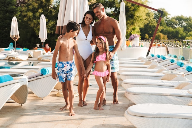 Foto de familia caucásica feliz con niños descansando cerca de la piscina de lujo con tumbonas y sombrillas de moda blanca, al aire libre durante la recreación