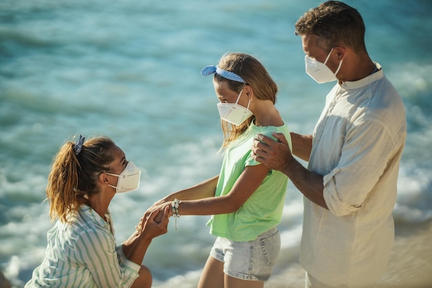 Foto de una familia feliz con máscara protectora N95 pasando tiempo en la orilla del mar en la pandemia de la corona.