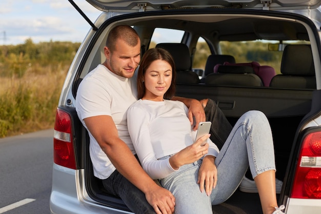 Foto de una familia joven y amorosa sentada en el baúl de un auto en la carretera, un hombre y una mujer con camisas blancas, haciendo selfie para encontrar el camino más corto mientras descansan usando el navegador del teléfono