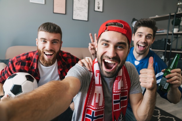 Foto de fanáticos de chicos emocionados jóvenes que muestran el pulgar hacia arriba y toman una foto selfie mientras ven un partido deportivo en la sala de estar