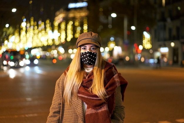 Foto fechada de uma jovem loira com uma máscara voltada para a câmera, em uma cidade à noite, com muitas luzes de fundo e carros passando. Atmosfera de inverno.