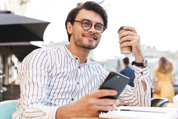 Foto de feliz hombre caucásico con anteojos bebiendo café y escribiendo en el teléfono móvil mientras trabajaba en la cafetería al aire libre