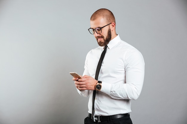 Foto de feliz hombre exitoso en gafas y corbata comunicarse mientras sostiene el teléfono inteligente en las manos, aislado sobre la pared gris