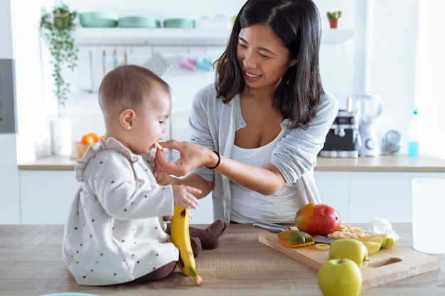 Foto de feliz joven madre alimentando a su linda niña con un plátano en la cocina de casa.