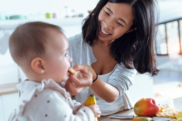 Foto foto de feliz joven madre alimentando a su linda niña con un plátano en la cocina de casa.