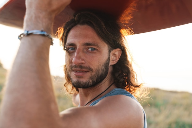 Foto de feliz joven sosteniendo la tabla de surf sobre su cabeza en la playa soleada