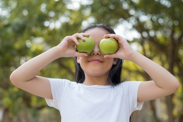 Foto feliz menina asiática em pé com um grande sorriso. segurando maçã verde em sua mão, bio natureza verde saudável fresca com folhagem borrada abstrata e luz do sol de verão brilhante