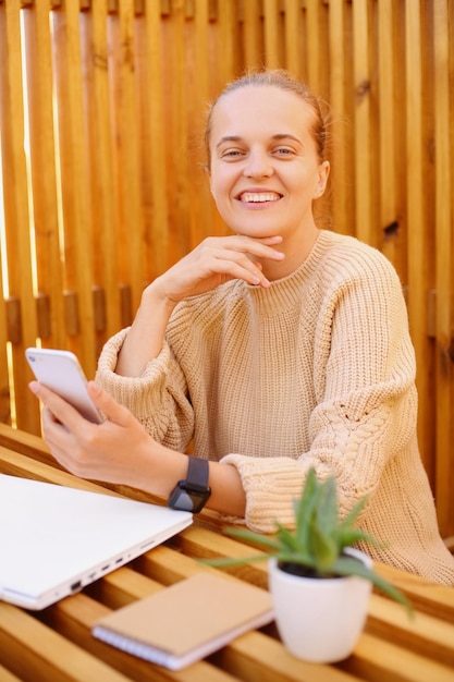 Foto de feliz y sonriente mujer caucásica feliz con peinado de moño usando suéter beige trabajando en una laptop y sosteniendo un teléfono inteligente en las manos mirando la cámara con expresión satisfecha