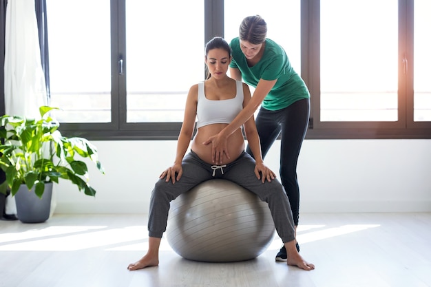 Foto de fisioterapeuta ayudando a hermosa mujer embarazada para hacer ejercicios de pilates con pelota preparándose para el parto.