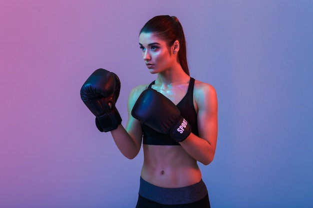 Foto de fitness saludable mujer de 20 años en boxeo de ropa deportiva en guantes negros durante el entrenamiento en el gimnasio, aislado sobre la pared púrpura