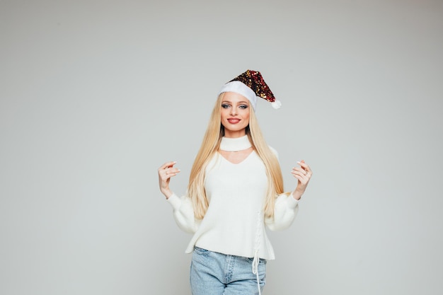 Foto genérica de la hermosa joven rubia en reluciente sombrero de Santa y blusa blanca y jeans azul claro sonriendo a la cámara sobre fondo blanco.