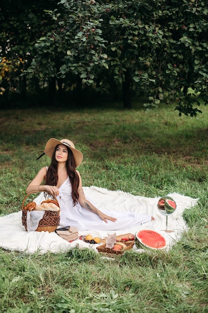 Foto genérica de longitud completa de una hermosa dama morena con sombrero de verano y vestido blanco sentada en la manta con una canasta de panadería y otros alimentos a su alrededor en la manta en el parque de verano.