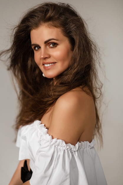 Foto genérica de la mujer caucásica bastante morena en blanco con el hombro desnudo sonriendo a la cámara. Mujer bonita con pelo ondulado ventoso con top blanco.