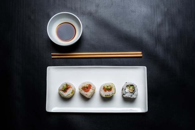 Foto genérica de sushi roll vegetal en plato de cerámica blanca, palillos y un tazón de salsa de soja visto desde arriba sobre un fondo negro.