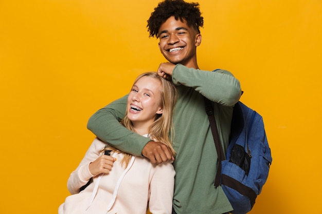 Foto de gente adolescente hombre y mujer de 16-18 con mochilas riendo y abrazándose juntos, aislado sobre fondo amarillo