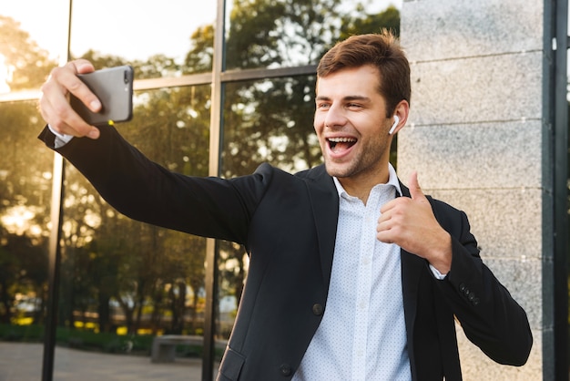 Foto de gerente ejecutivo en traje sosteniendo teléfono móvil para selfie, mientras está parado al aire libre contra el edificio