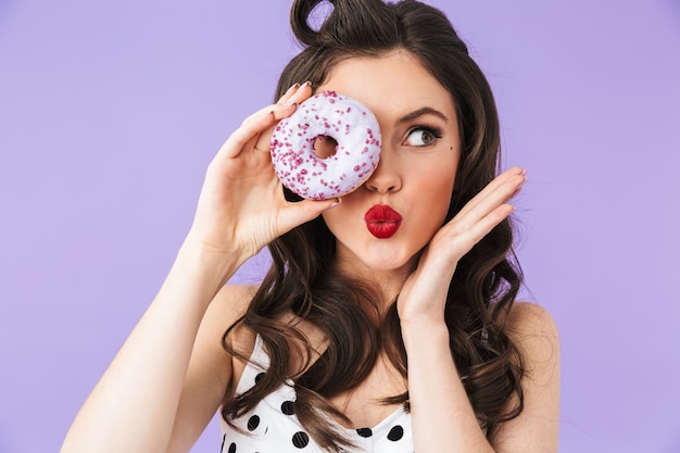 Foto de glamour pin-up mujer en vestido de lunares vintage sonriendo mientras sostiene y come sabroso donut aislado sobre pared violeta