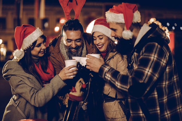 Foto de un grupo de alegres jóvenes amigos divirtiéndose juntos y disfrutando del vino caliente en el mercado navideño en una fiesta nocturna.