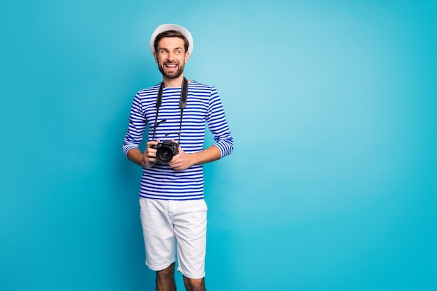 Foto de guapo chico guapo turista mantenga cámara de lente digital profesional viajando al extranjero mirar espacio vacío usar camisa de marinero a rayas gorra pantalones cortos color azul aislado