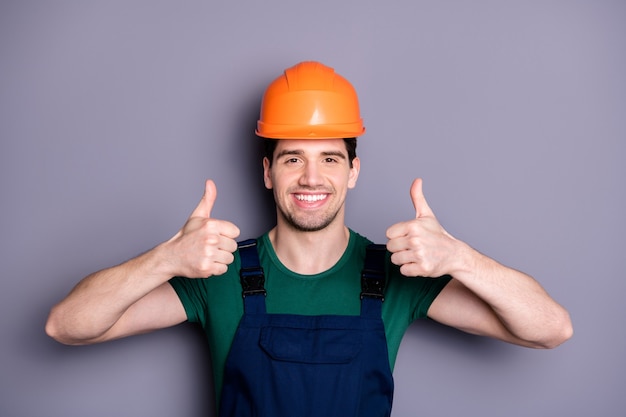 Foto de guapo trabajador masculino ingeniero calificado levantar los dedos pulgar hacia arriba aconsejando buena calidad usar camiseta azul mono de seguridad casco protector aislado pared gris