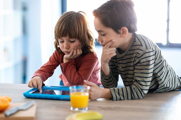 Foto de hermanos jóvenes aburridos jugando con la tableta digital mientras está sentado en la cocina de su casa.