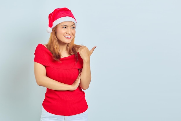 Foto de hermosa chica asiática con sombrero de navidad con expresión facial sonriente y apuntando a un lado sobre fondo blanco.