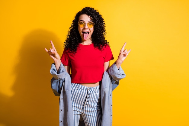 Foto de hermosa dama con lengua fuera mostrar cuernos usar camiseta roja chaqueta de jeans a rayas fondo de color amarillo aislado