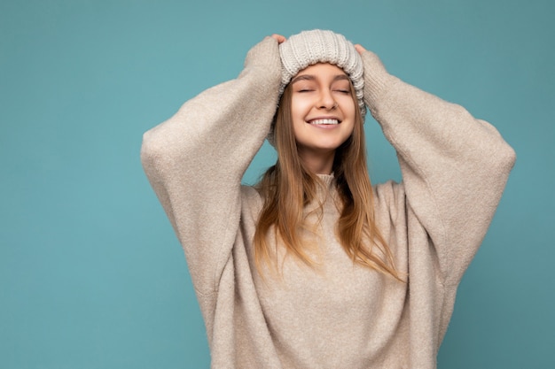 Foto de hermosa feliz alegre joven rubia oscura persona femenina aislada sobre fondo azul pared vistiendo suéter beige cálido y sombrero beige tejido disfrutando con los ojos cerrados. Copia espacio