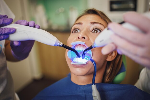 La foto de una hermosa joven está en el dentista. Ella se sienta en la silla del dentista y el dentista fijó cerraduras linguales en sus dientes con luz de curado.