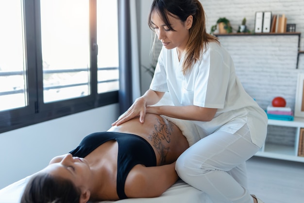 Foto de hermosa joven fisioterapeuta masajeando la barriga de la mujer embarazada en una camilla en casa.