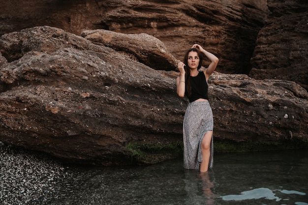 Una foto de una hermosa joven sensual con cabello largo y oscuro en un vestido posando en la playa junto a grandes rocas