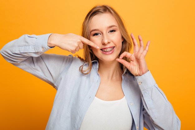 Foto de hermosa mujer de 20 años con cabello castaño sonriendo y gesticulando con un signo bien mientras señala con el dedo los frenos en los dientes, aislado sobre fondo amarillo