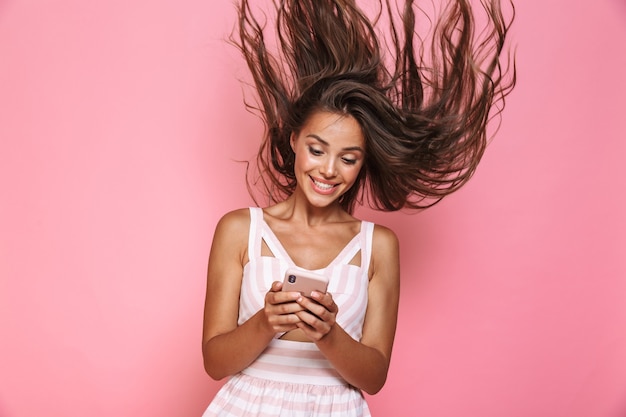 Foto de hermosa mujer de 20 años vistiendo un vestido sonriendo y sosteniendo el teléfono móvil con el pelo temblando, aislado sobre la pared rosa