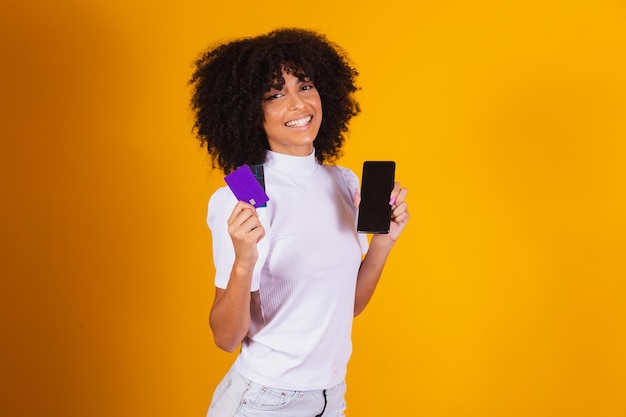 Foto de una hermosa mujer emocionada de piel oscura que sostiene una tarjeta de crédito de plástico que muestra un nuevo modelo de teléfono que aconseja el pago en línea con fondo de color amarillo