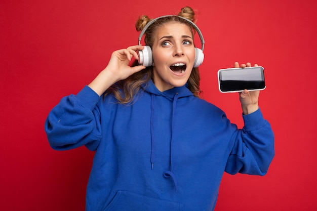 Foto de hermosa mujer joven divertida feliz positiva vistiendo elegante sudadera con capucha azul aislado sobre rojo