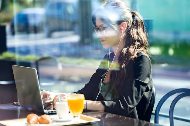 Foto de hermosa mujer joven que trabaja con su computadora portátil mientras desayuna en una cafetería.