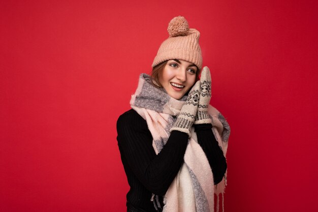 Foto de hermosa mujer morena joven linda feliz aislada sobre la pared de fondo rojo con guantes de bufanda de invierno y gorro mirando hacia el lado y soñando.