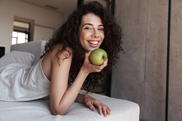 Foto de hermosa mujer rizada con cabello largo y oscuro con ropa de ocio de seda comiendo manzana verde fresca con placer y sonrisa, mientras está acostado en el sofá en casa