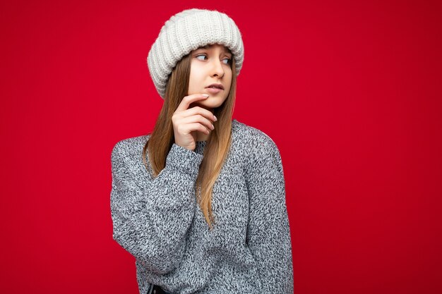 Foto de hermosa mujer rubia adulta preocupada triste pensativa que se encuentran aisladas sobre la pared de fondo rojo vistiendo un suéter gris y un sombrero beige mirando hacia el lado. Espacio vacío, espacio de copia