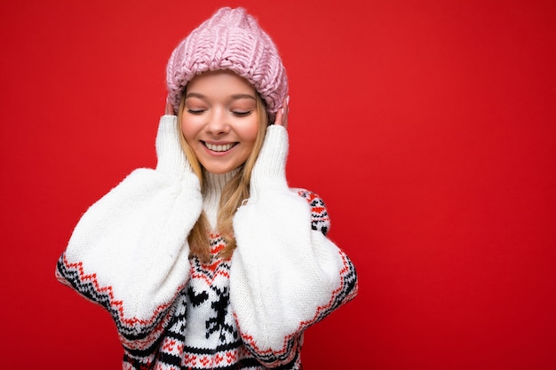 Foto de hermosa mujer rubia joven feliz aislada sobre fondo rojo pared vistiendo suéter de invierno y sombrero rosa mirando hacia abajo y divirtiéndose. Espacio vacío, espacio de copia