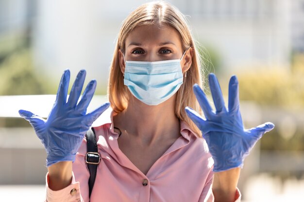 Foto de una hermosa mujer rubia con máscara protectora y guantes mostrando las manos a la cámara en la calle.