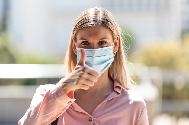 Foto de hermosa mujer rubia en mascarilla mirando a cámara y mostrando el pulgar hacia arriba en las calles de la ciudad.