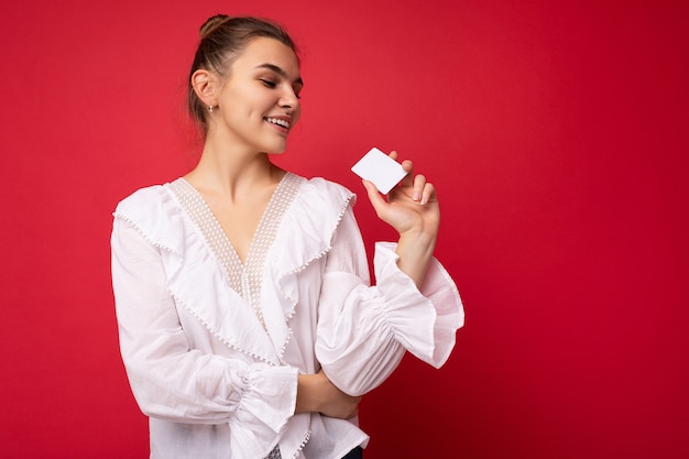 Foto de hermosa mujer rubia oscura joven feliz con blusa blanca aislada sobre pared roja con tarjeta de crédito mirando tarjeta de plástico. espacio vacio