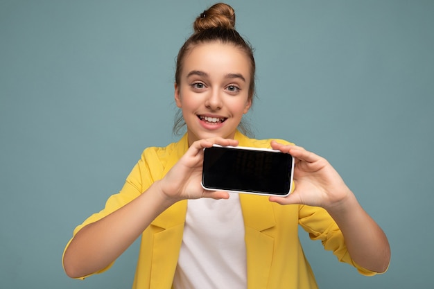 Foto de hermosa niña sonriente guapa vistiendo traje casual elegante de pie aislado en el fondo con espacio de copia sosteniendo teléfono inteligente mostrando el teléfono en la mano con pantalla vacía para maqueta