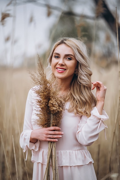 Foto de una hermosa niña sonriente con el pelo largo y rubio y rizado en vestidos largos y claros de pie en un campo de caña