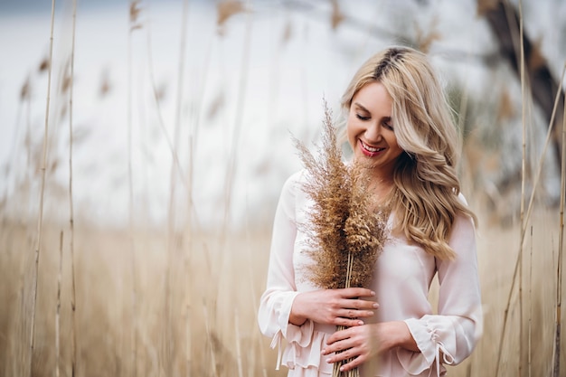 Foto de una hermosa niña sonriente con el pelo largo y rubio y rizado en vestidos largos y claros de pie en un campo de caña