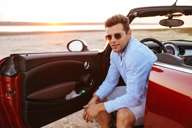 Foto de hermoso hombre caucásico con gafas de sol sentado en un elegante coche convertible junto al mar al amanecer.