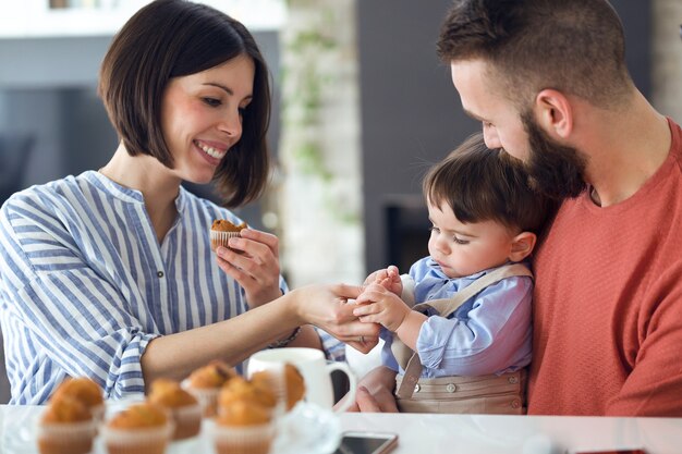 Foto de hermosos padres jóvenes con su bebé comiendo muffins en casa.