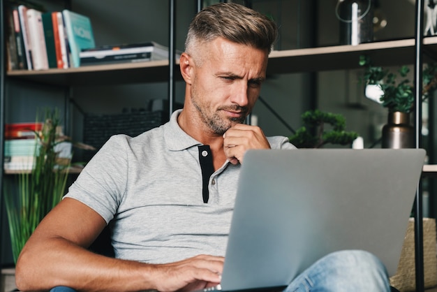 Foto de hombre adulto seguro en ropa casual usando laptop mientras está sentado en un sillón en el apartamento